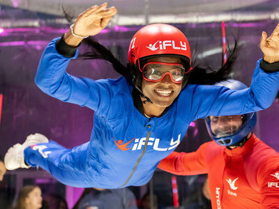 a woman wearing a blue flight suit, goggles and helmet being helped by an instructor in a red suit on an indoor skydiving experience