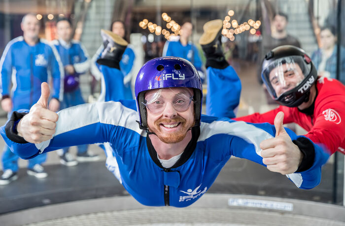 a man wearing a blue flight suit, goggles and helmet giving the thumbs up on an indoor skydiving experience